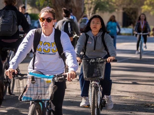 a closeup on two students riding among other students down a road on campus.