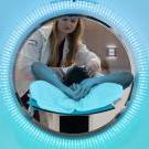 在一个圆形的开口, 一位身穿白色实验室大褂的长发女子俯身看着一个躺着的人，他们的头顶正对着摄像机. 蓝色填充圆圈外的其余区域. 