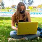澳门十大电子游戏大全游泳池前的草地上，一名学生戴着面具在她的笔记本电脑上工作.